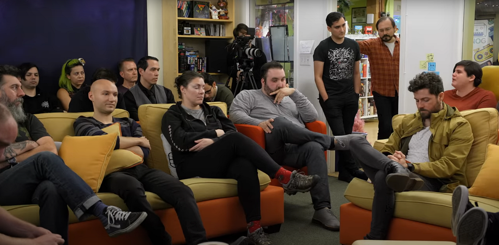 Double Fine employees sit on couches during an uncomfortable meeting. Many nervously cross their arms and avert their gaze to the floor. On the right, one employee is in the middle of an impassioned comment