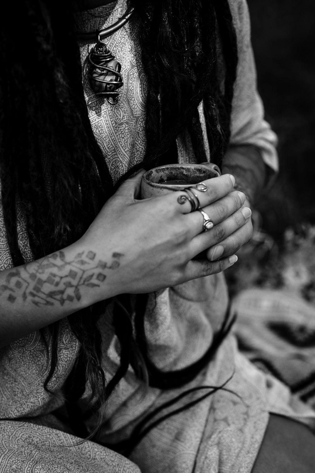 A black and white photo of a close-up of part of a person’s torso and their clasped hands holding what appears to be a small cup. The person has long dark dreadlocks, a tattooed pattern on their arm, and is wearing a light patterned garment, a necklace with a pendant bound in wire, and two finger rings.