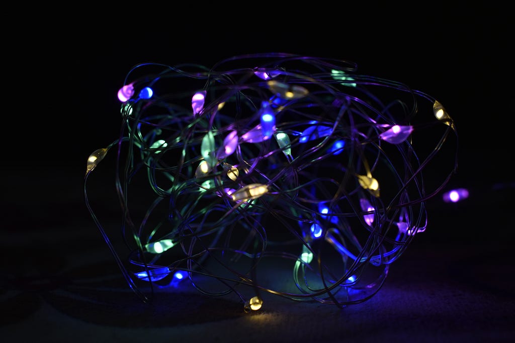 Photo of a tangled string of Christmas lights in the dark, with the small pools of of multicoloured light illuminating the wires that connect them.