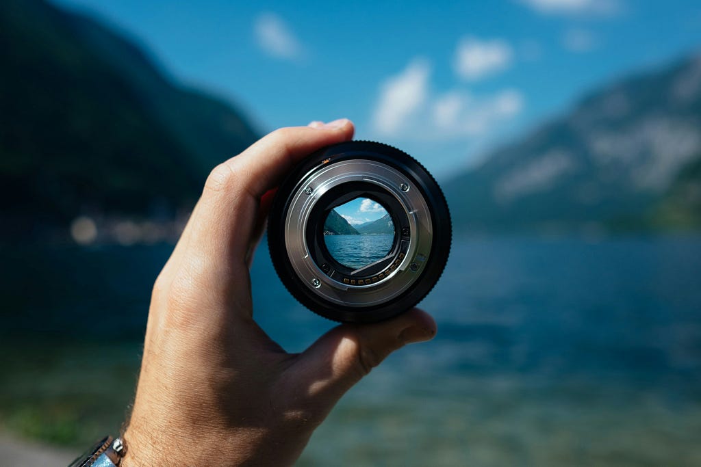 A hand holding up the lens of a camera which captures the distant view of a mountain and lake