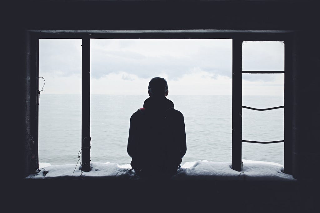 a lone man, sitting on a windowsill, facing the ocean, seen from behind.