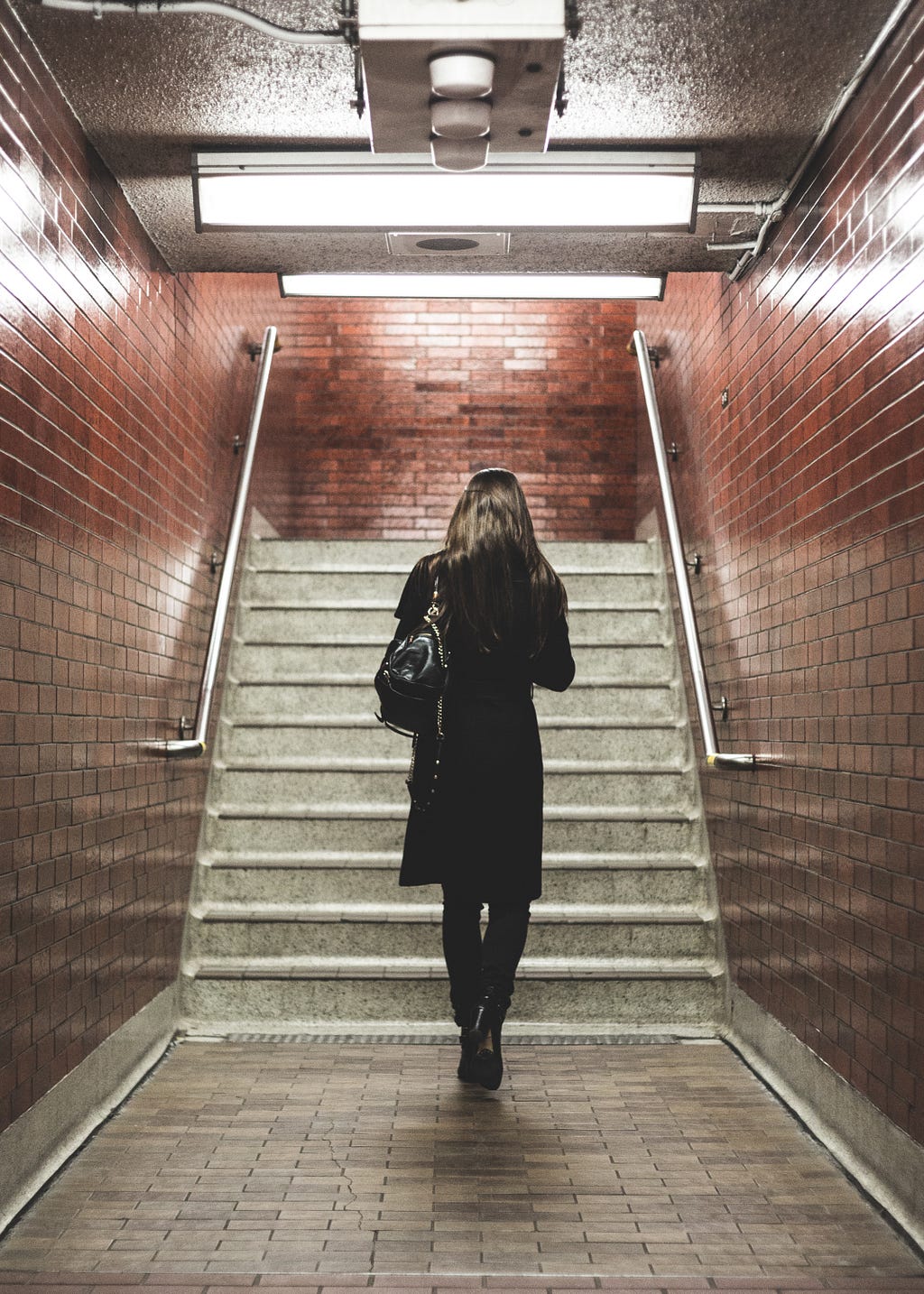 Image of a woman walking away, long hair, black outfit, black purse, heading towards stairs. This might be an underground subway or train station.