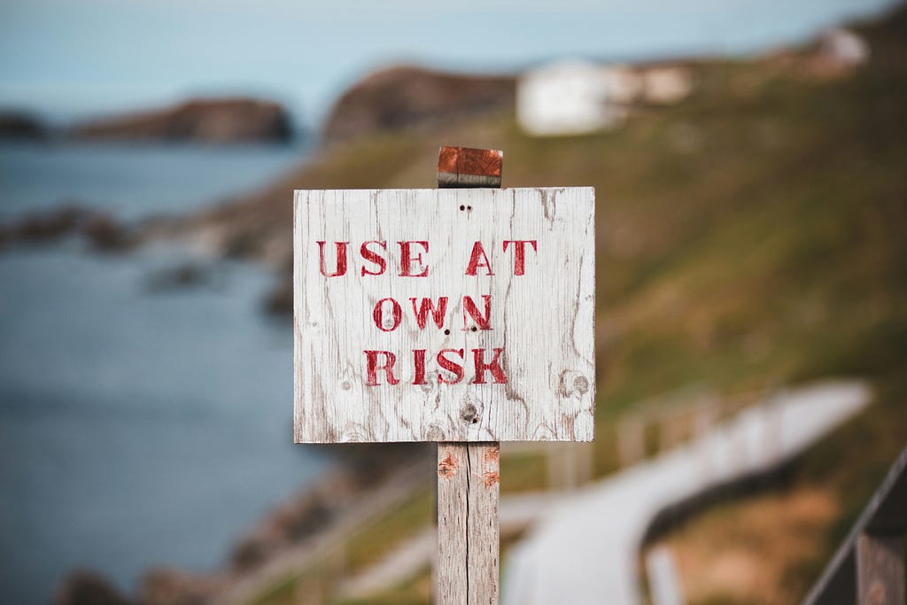 A simple wooden sign against an out of focus coastal backdrop. The sign is white and in all-caps red lettering reads “USE AT OWN RISK”