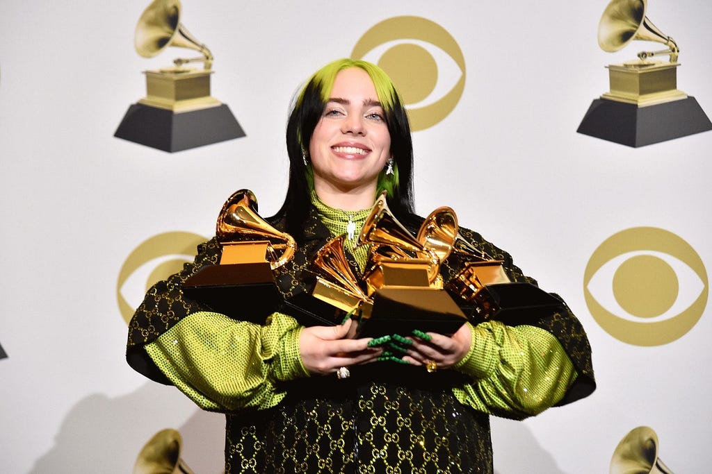 Eilish stands smiling, holding the five grammys she won at the 2020 Grammy Awards