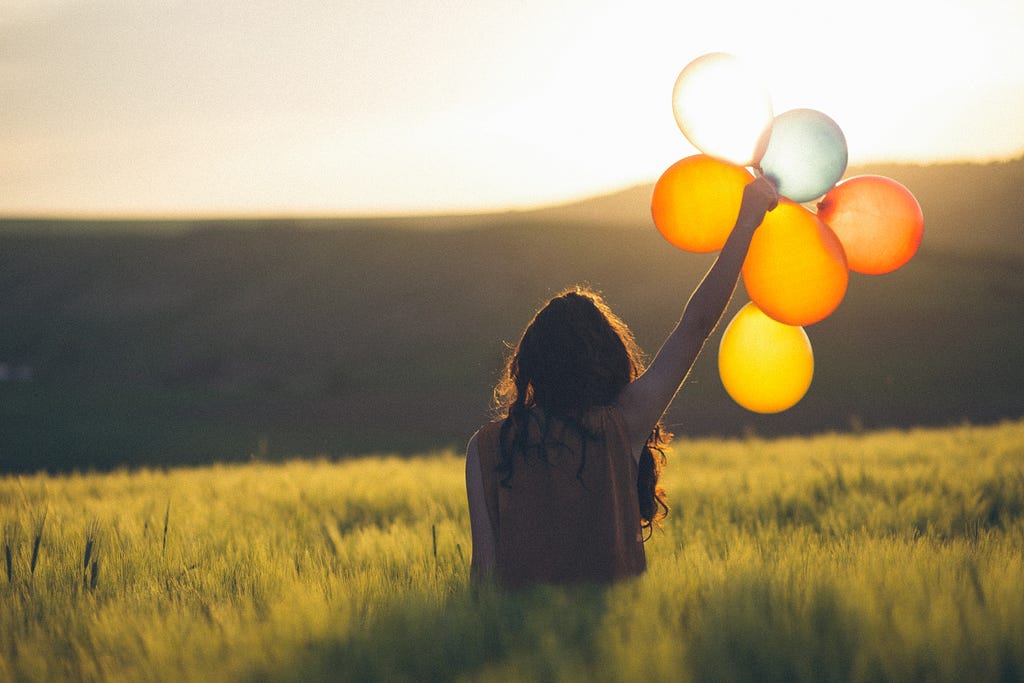 A person with long hair shown from behind holding balloons in a field where the sun shines through and illuminates them