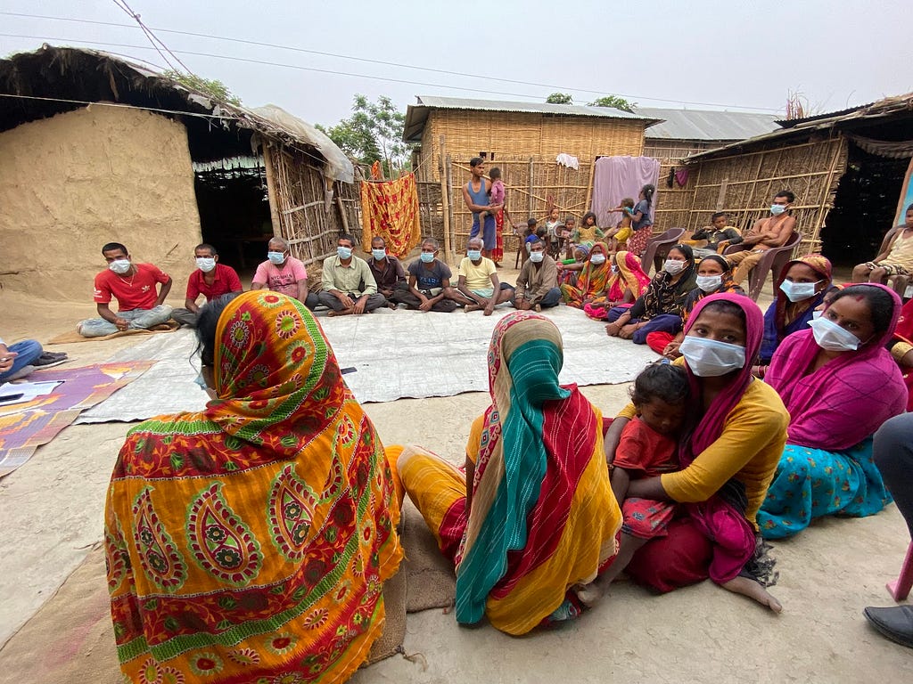 Community members sit cross-legged in a large semicircle wearing colorful clothing and protective face masks. One woman looks towards the camera holding her child.