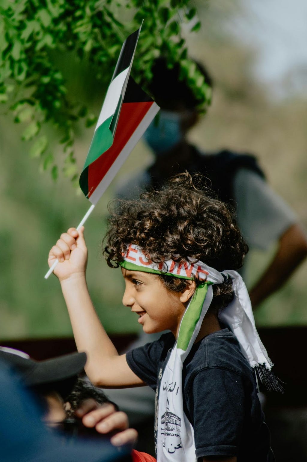 A boy in blue and white shirt holding a Paslestine flag during the daytime.