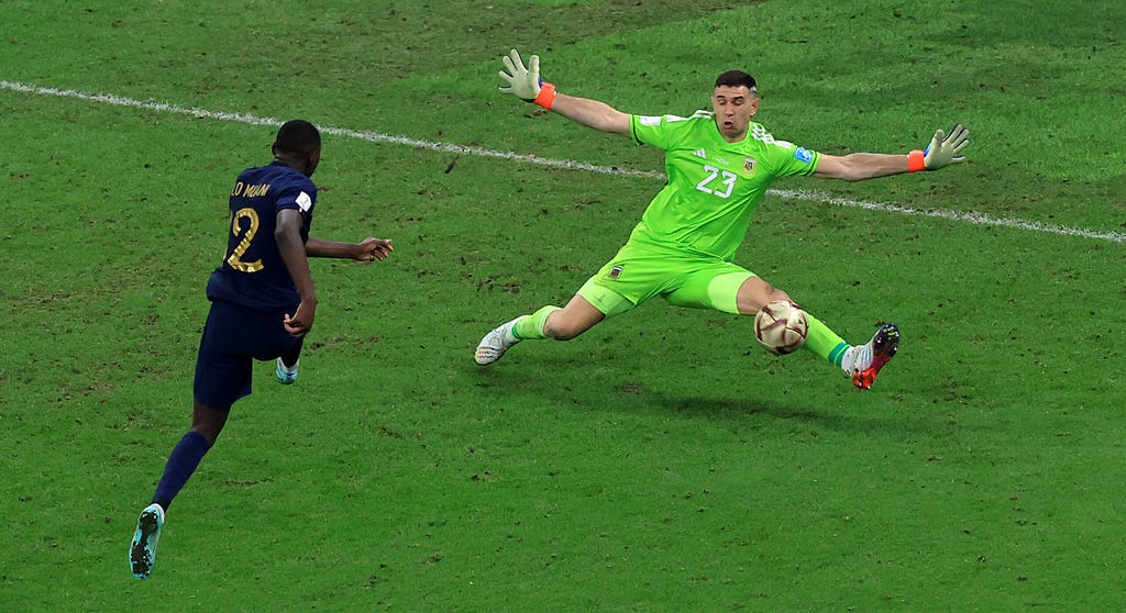 O camisa 12 da França chuta a bola em dieração ao gol no último lance da porrogação, e o goleiro argentino Martinez defende com o pé