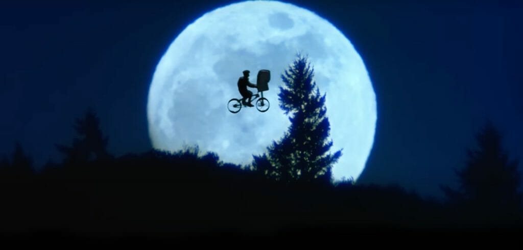 Elliot and E.T flying on a bike over a dark forest, and a bright round moon in the background.