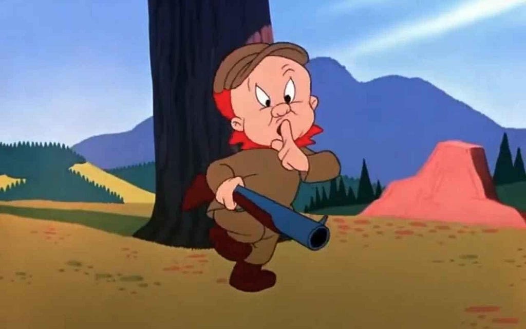 A still from the theatrical cartoon short “Rabbit Fire” (1951), showing the hunter Elmer Fudd in his usual brown hunting clothes, looking at the audience and holding his finger up in a ‘shh’ gesture. He is holding a rifle.