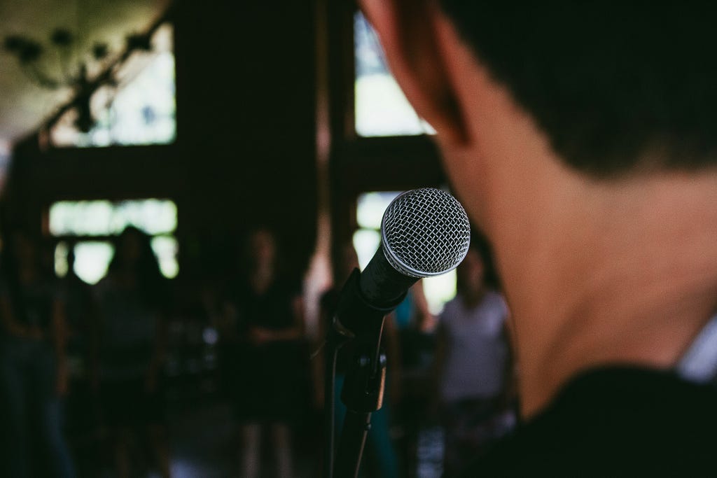 The back of a mans head as he stands in front of a microphone, the people in the background are blurry putting the emphasis on the speaker.