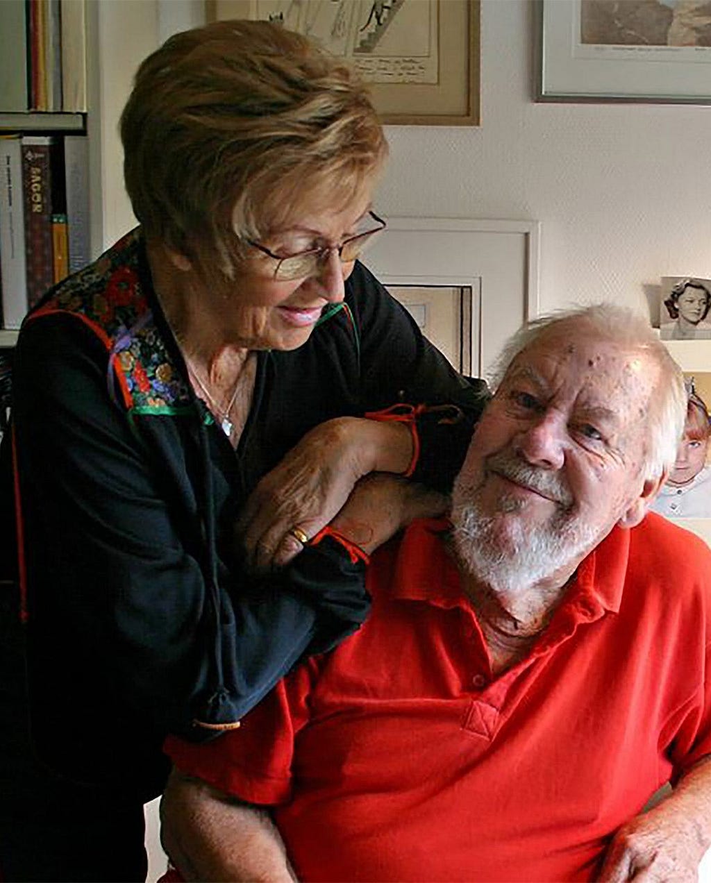 An older woman with short-cropped hair and wearing glasses is leaning tenderly on the shoulder of an older man seated in a chair. She smiles gently downwards, wearing a black jacket with colorful embroidery, and her arms are folded comfortably around the man’s shoulder. The man, with a full white beard and bright eyes, is wearing a red polo shirt and smiling broadly at the camera. They are in a room filled with cultural artifacts: a bookshelf with neatly organized books to the left and multiple