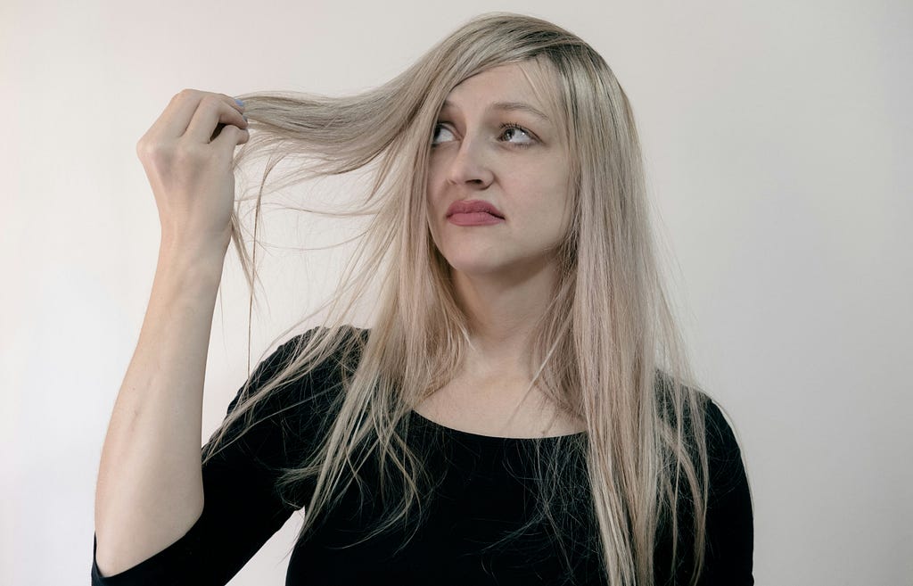 A 30-something Caucasian woman with long blonde hair is looking up to her right, holding the end of her hair, with a disapproving look on her face.