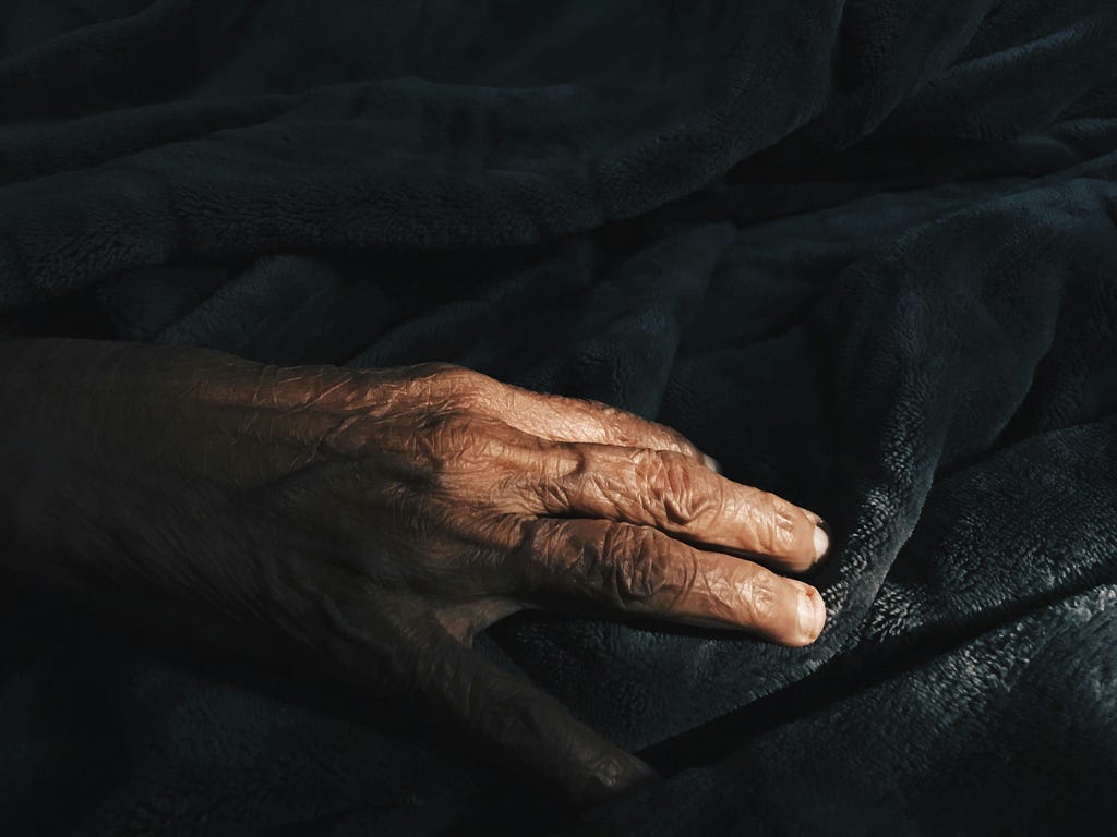 An image of an elderly Black person’s hand resting on a black background. A beam of light shines across their fingers.