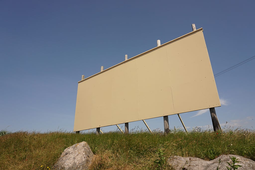 Middle-distance, low-angle, daytime photograph of a blank, 4-post billboard, in a grass field w/rocks in the foreground.