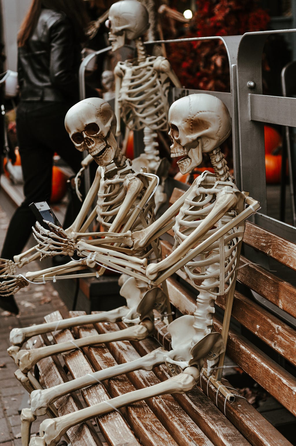 2 skeletons sit on a city bench, one is holding a smart phone