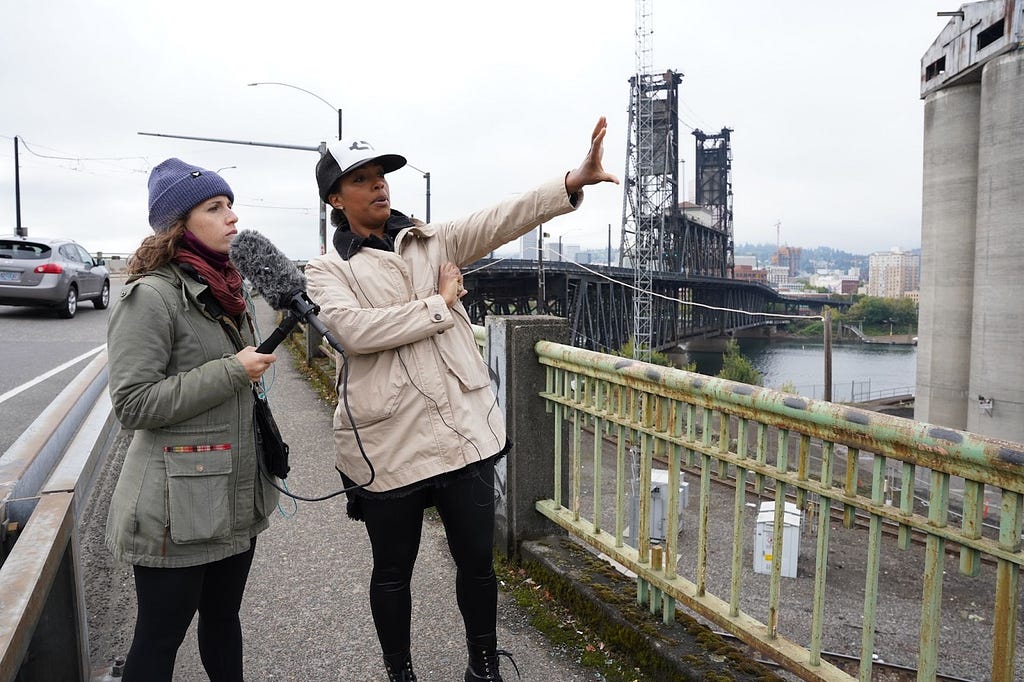 Photograph of two women, one holding a microphone, standing on a bridge’s sidewalk, between the road and an industrial lot.