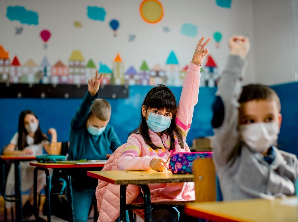 Kids wearing face masks raising their hands in an elementary school classroom. Photo by kevajefimija/Getty Images