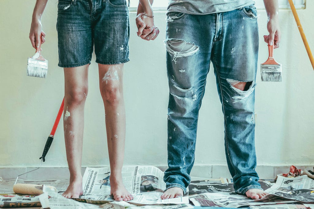 A couple holds each other’s hands and grasp paintbrushes in the other. They’re about to paint a wall in their home.