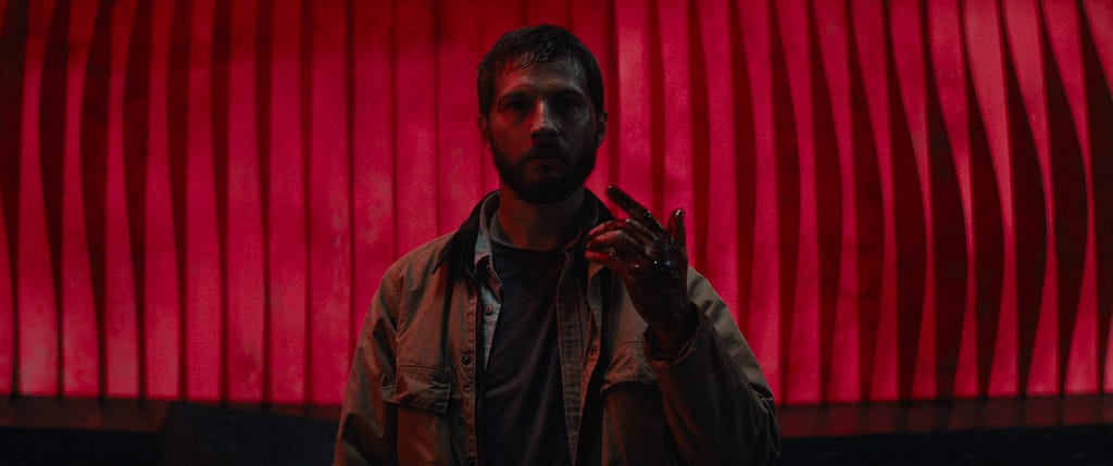 A still from Upgrade (2018) showing a bloodied man, Grey Trace (Logan Marshall-Green), standing in front of a red, wave-patterned wall, holding up his hand as if realizing he’s no longer in control.