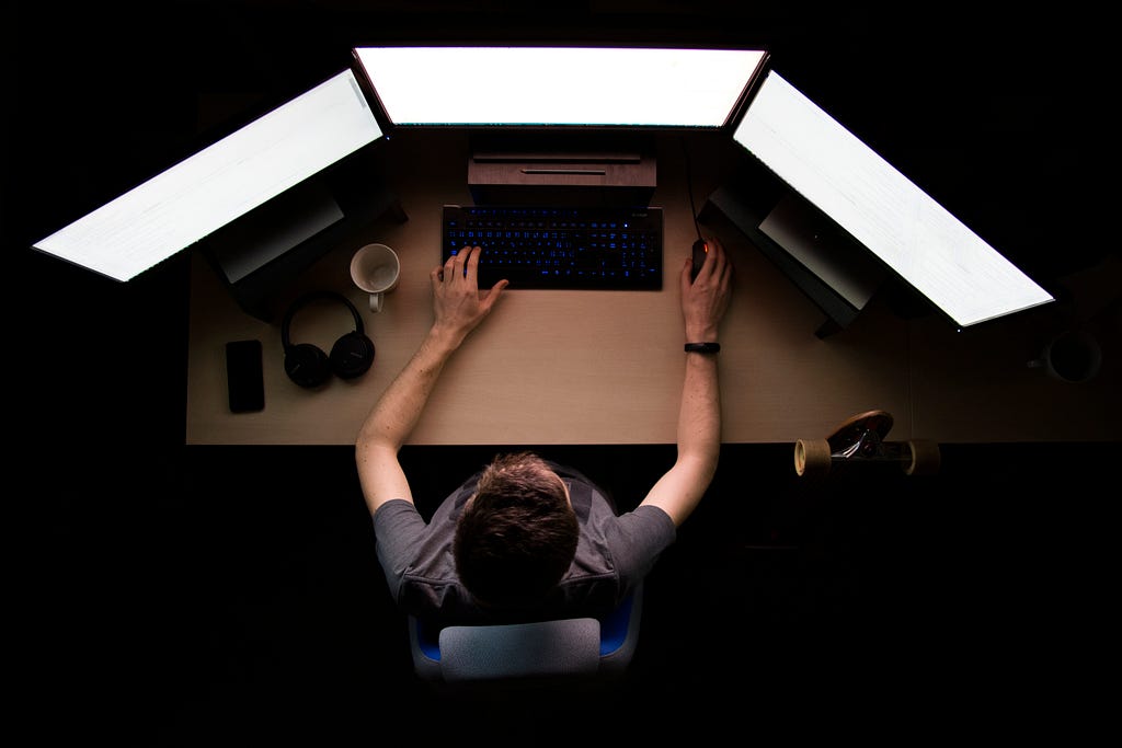 A man staring at a computer. He has a three-monitor setup, drinks cofee and maitains his phone and headset nearby.