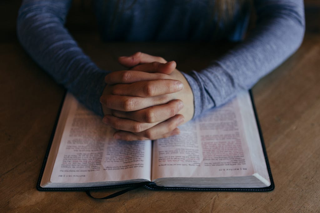 a person is praying to God with their hands together and an open bible underneath. they are talking to God, building a relationship with Him.