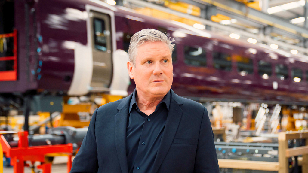 UK PM Keir Starmer wearing a Black Suit standing in front of a train being build in a factory via the Labour Party.