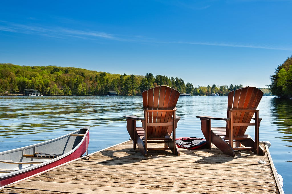 Two Muskoka chairs on a wooden dock overlooking the blue water of a lake in Muskoka, Ontario Canada.