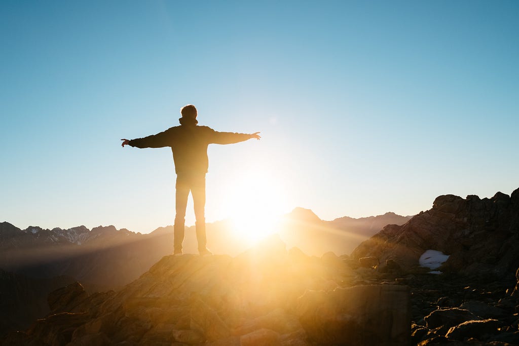 A mountain climber looking out at the rising sun among the peaks of mountains.