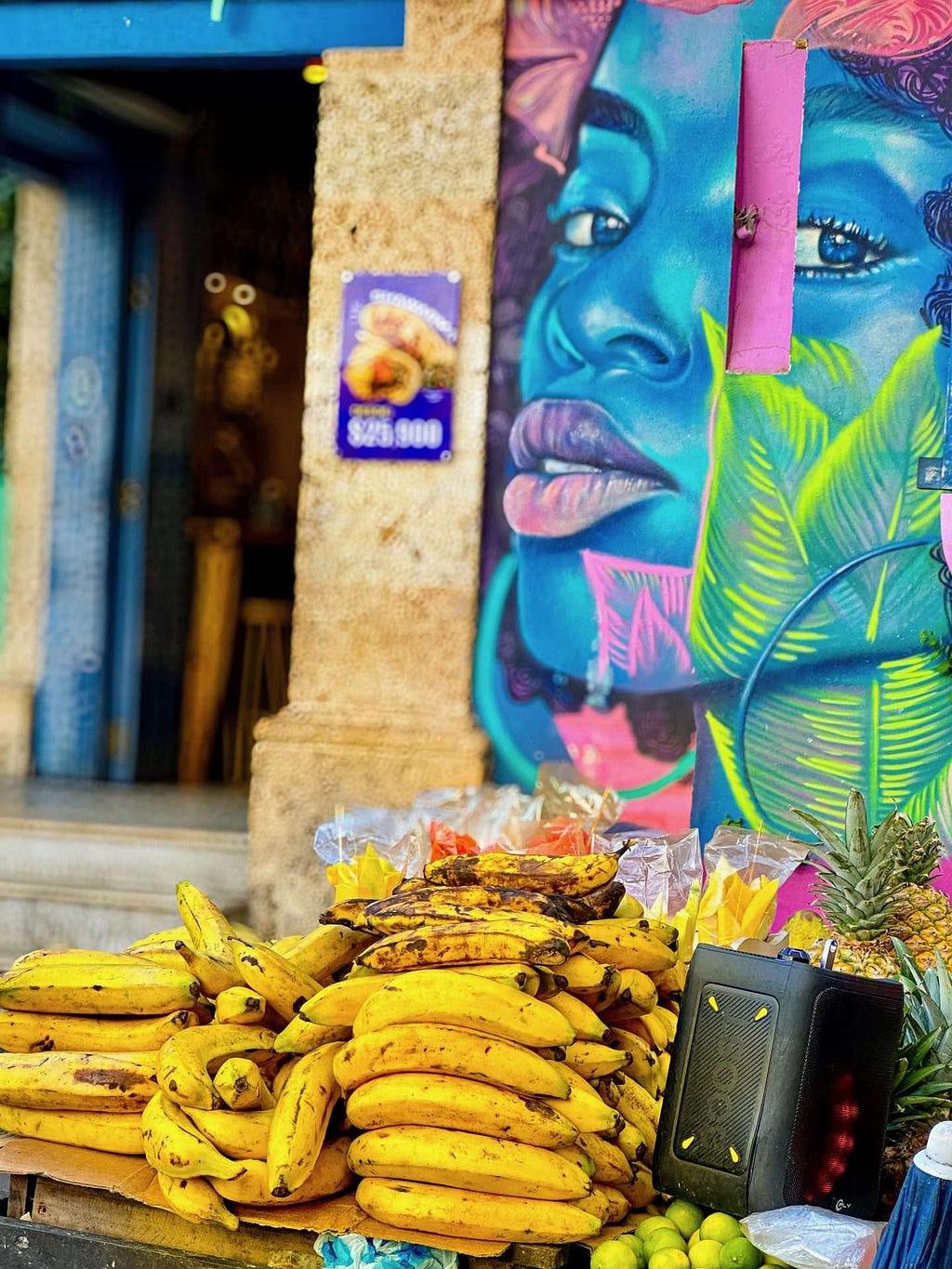 A pile of ripe bananas and other fruits are on display at an outdoor market stand in the neighborhood of Getsamani in Cartegena, Colombia. In the background, there is a colorful mural of a woman's face.