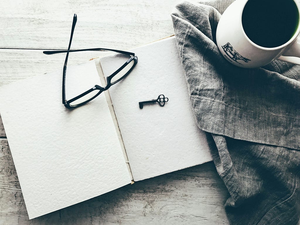 Black and white photo of a wooden table on which, from right to left, there is a cup with what looks like coffee, on a denim placemat, an open journal with white sheets, on top of it a pair of glasses and a key.