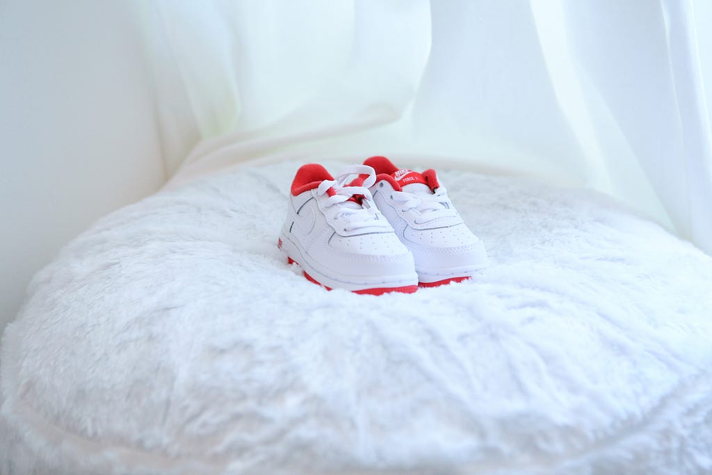 White baby shoes with a red outline sit on a perfectly made fluffy white bed.