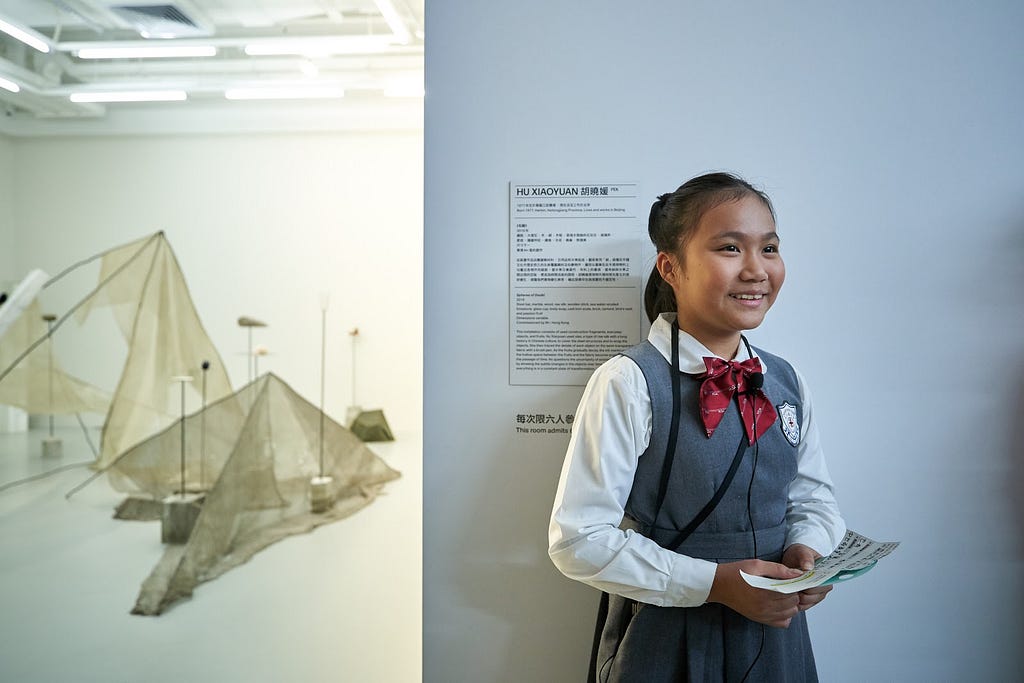 A young student stands smiling inside a gallery space, next to the opening to a room containing an installation artwork.