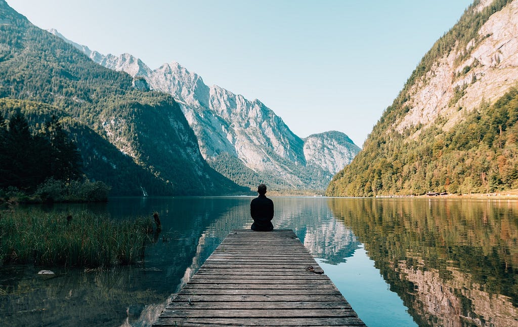 Serene picture of a person sitting at the end of a dock looking over a beautiful lake with mountains in the background