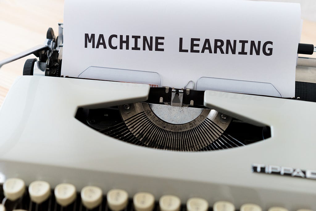 This is a photo of a typewriter with a paper in it. The paper reads “MACHINE LEARNING” in all caps. The typewriter is a light beige color with white keys. The typewriter is on a surface. The background is blurred and appears to be a light colored wall.