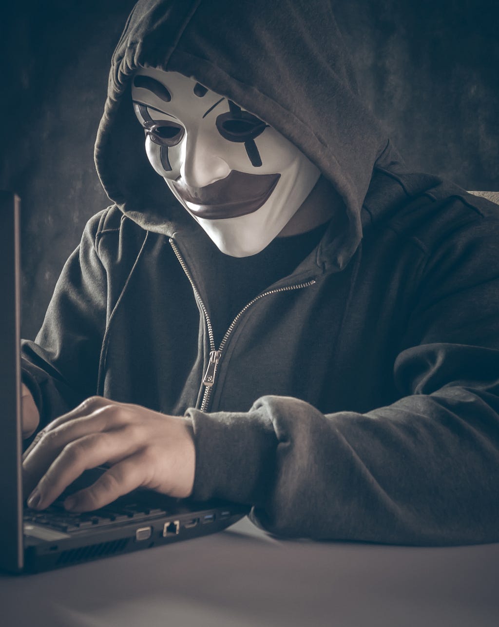 Hooded figure wearing a ‘Joker’ mask typing in front of a computer.
