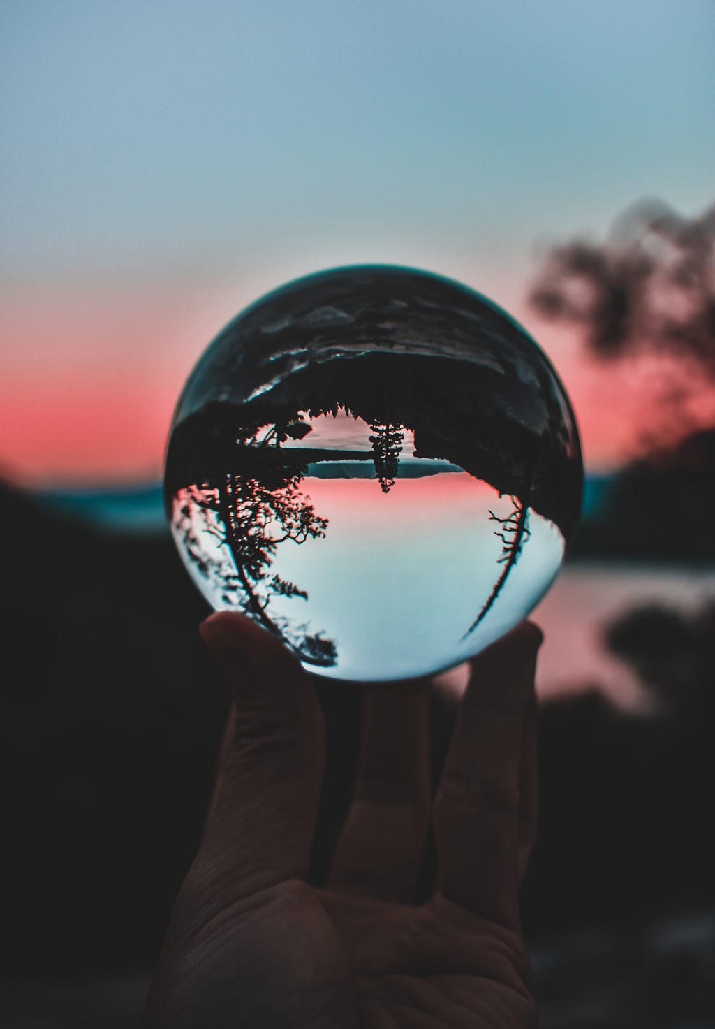 A crystal clear glass ball being held in front of a dusky pink, wooded, and mountainous lake landscape. The background image of the landscape is out of focus, and the inverted+spherically warped image of the same landscape can be seen in focus within the foreground image of the glass ball.