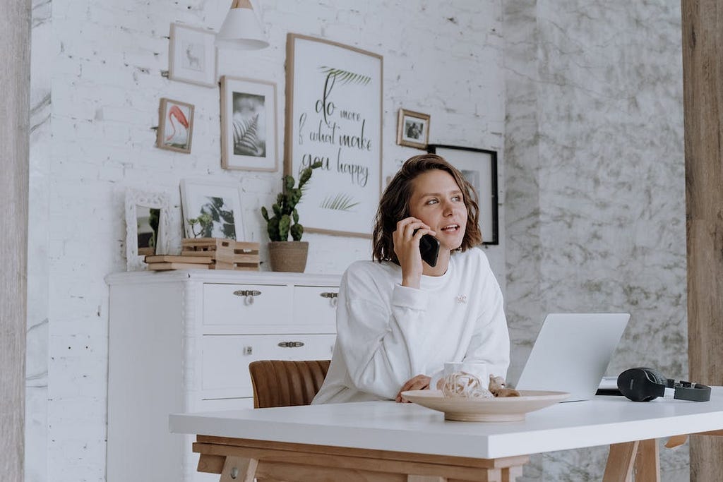 A woman in a white robe is talking on a phone call while working on her laptop at her desk. cottonbro/Pexels.