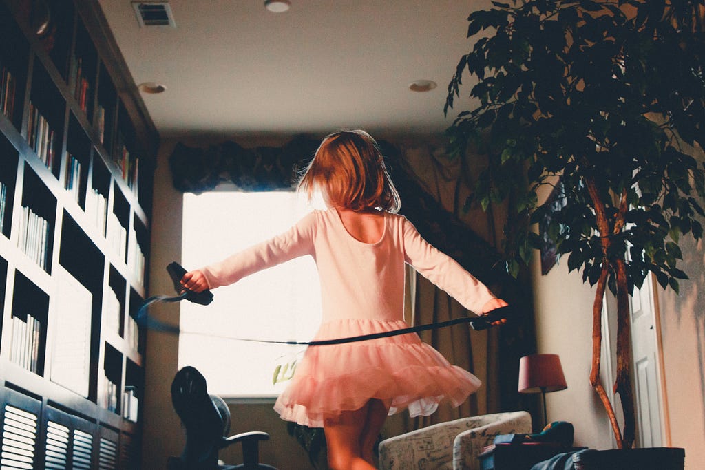 A child in a pink long-sleeved bodysuit and tutu plays with an exercise band in her living room.