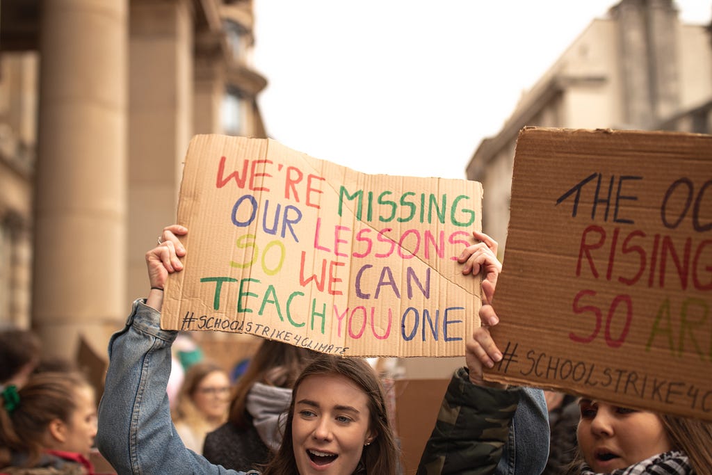 A young student holds a sign that says “we’re missing our lessons so we can teach you one #schoolstrike4climate” in multicolor.