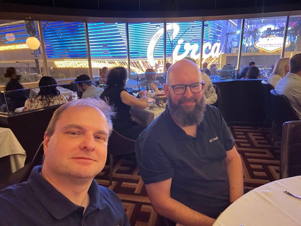 Two men are sitting at a table inside a bustling restaurant with a large illuminated Circa sign visible through the window behind them.