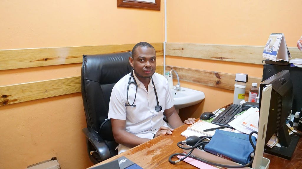 A doctor with a stethoscope draped across his shoulders sits behind his desk in his office.