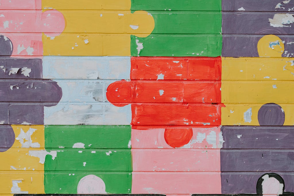 A faded and peeling painting on a wall made of wooden beams. The painting is of multicoloured jigsaw pieces fitted together