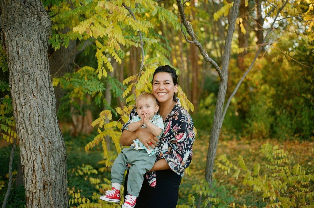 Portrait of Renee holding her child. She has dark hair, is wearing a printed shirt, and smiling at the camera. She holds a blonde child wearing a patterned shirt, green pants, and bright red shoes. They are standing in front of a forested background.