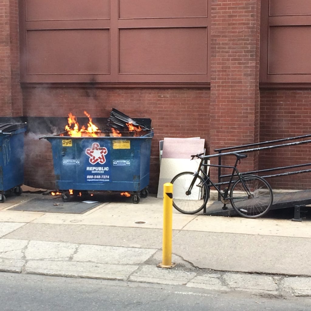 On a city sidewalk is a blue dumpster, bright orange flames peeking out.