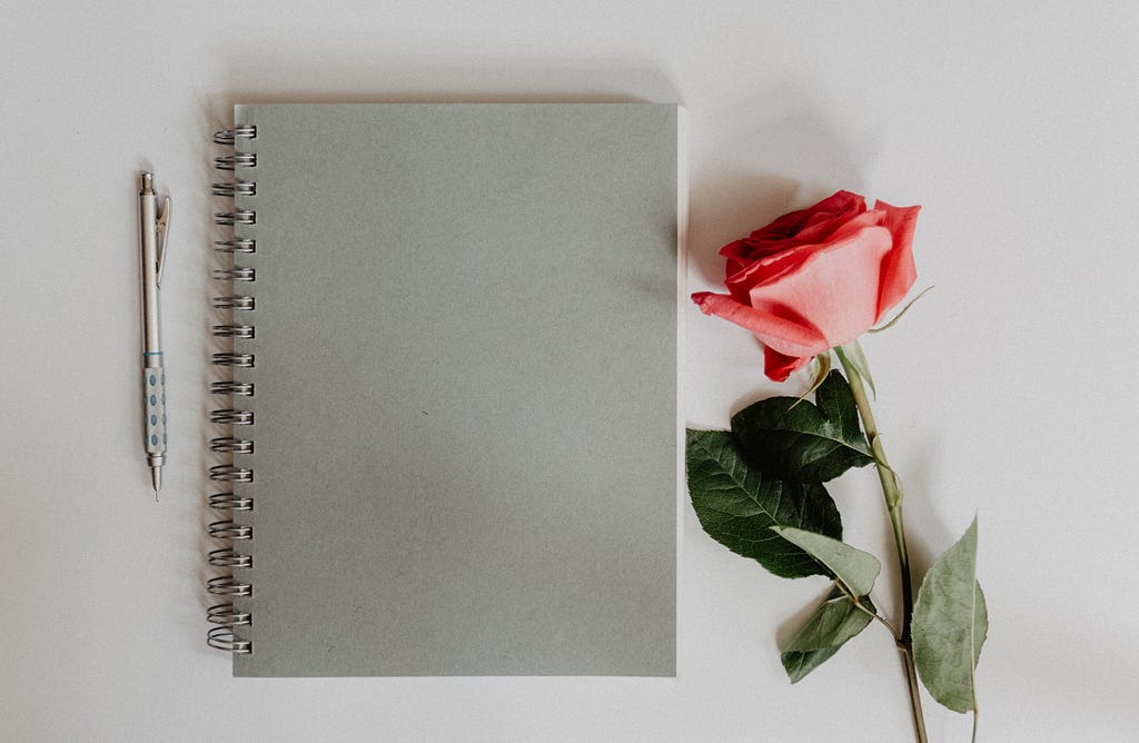 A grey notebook and pen, and a large, blooming red rose resting on its side, all sit against a grey-ish white background.