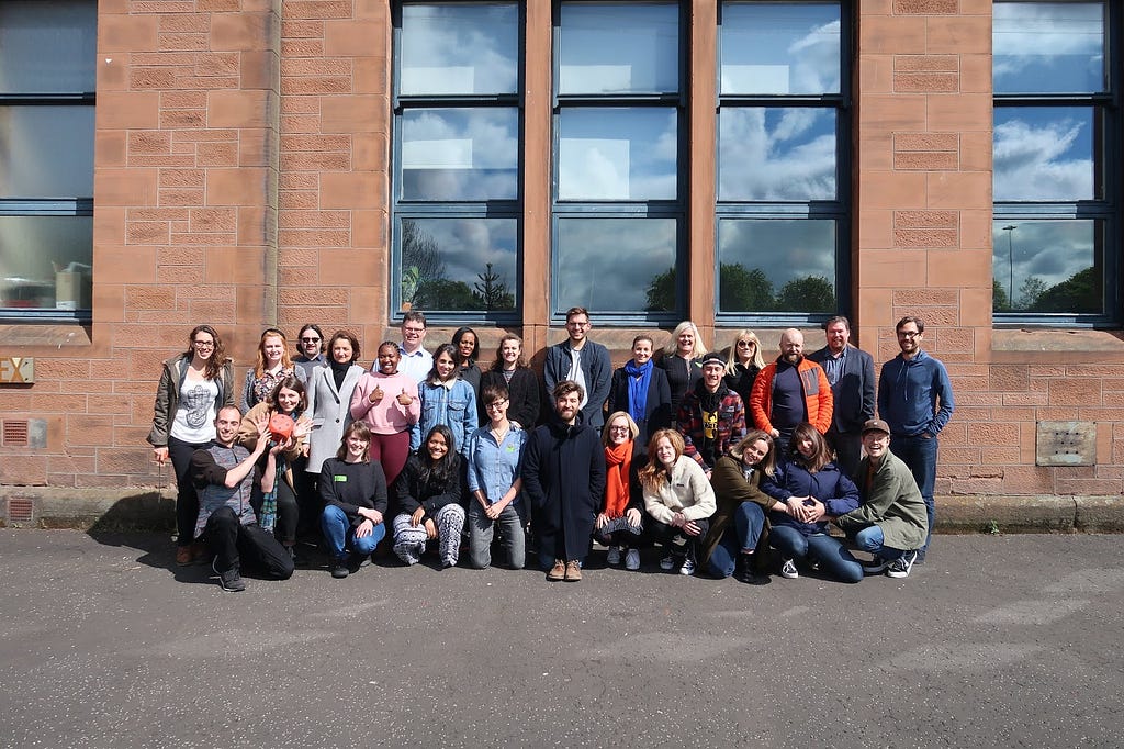 The Snook team, about 30 people standing in front of a brown wall with large glass vertical windows