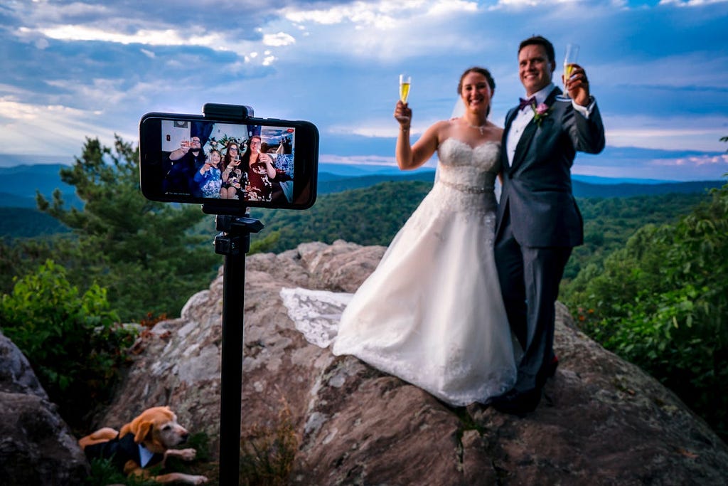 A bride and groom on a mountain give a virtual cheers.