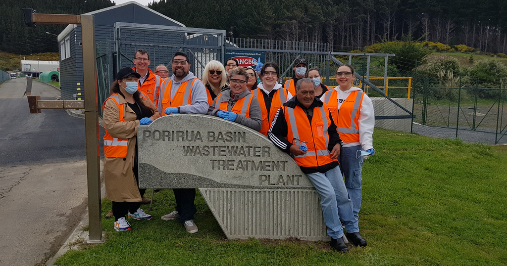 Image description: A group of mana whenua from Ngāti Toa wearing high vis vests are standing around the sign for Porirua Basin Wastewater Treatment Plant. The fence and one of the buildings of the waste treatment plant are in the background.