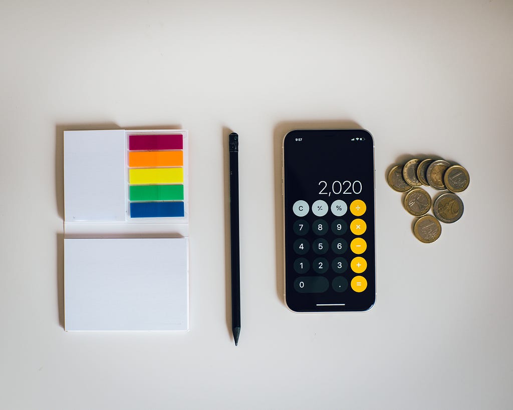 A phone with the calculator app open alongside some coins, small notepad and a pen